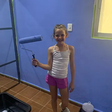 Young girl holding paintbrush in front of blue wall during art activity indoors.