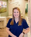 Woman in blue scrubs smiling at camera in modern dental office setting.