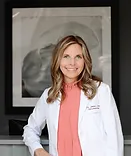Professional woman in white blazer and pink blouse smiling at camera, gray background, medical office setting.