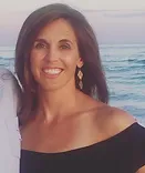 Woman smiling at camera with ocean backdrop, wearing off-shoulder black top