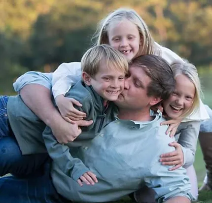 Man hugging four smiling children outdoors in natural daylight setting.