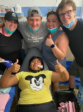 Four adults and one child in yellow shirt smile together indoors, showing thumbs up in casual group photo.